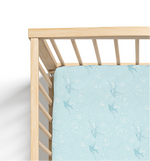 A crib sheet with a light blue background featuring a pattern of blue sparrows in flight and cloud-like designs, fitted on a standard crib.