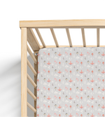 A crib sheet with a pattern of various stylized mushrooms in colors beige, gray, brown, and pink on a light gray background, fitted on a crib.