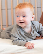 A smiling baby with red hair lies on their stomach on a white mattress inside a light wood crib, wearing a gray long-sleeve romper.