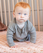 A baby with red hair and blue eyes wearing a gray long-sleeve henley shirt, sitting on a patterned pink sheet in a wooden crib.