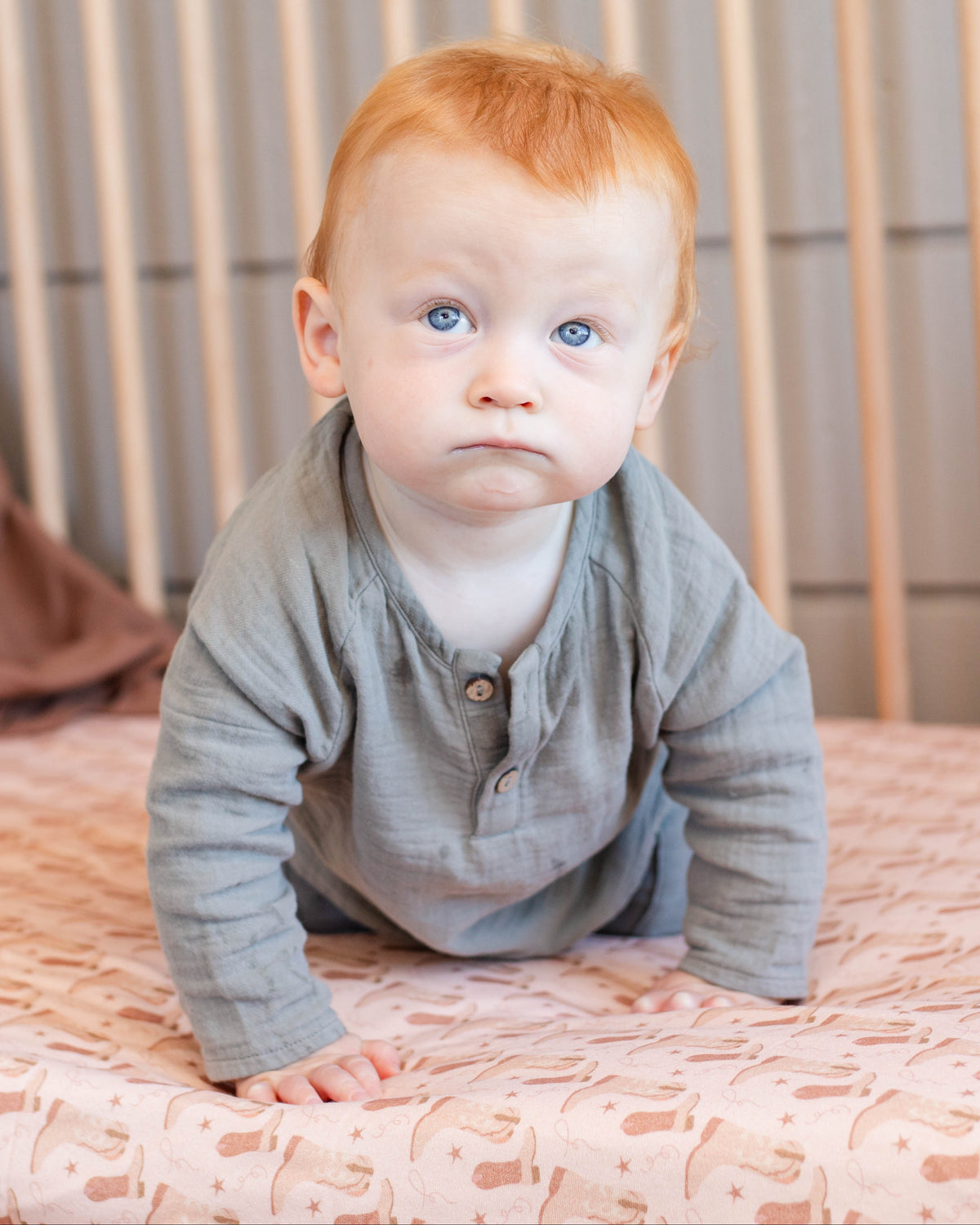 A baby with red hair and blue eyes wearing a gray long-sleeve henley shirt, sitting on a patterned pink sheet in a wooden crib.