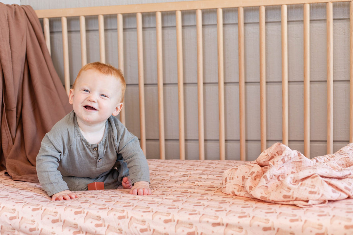 A smiling baby wearing a gray long-sleeve henley shirt sits on a pink patterned crib mattress inside a wooden crib.