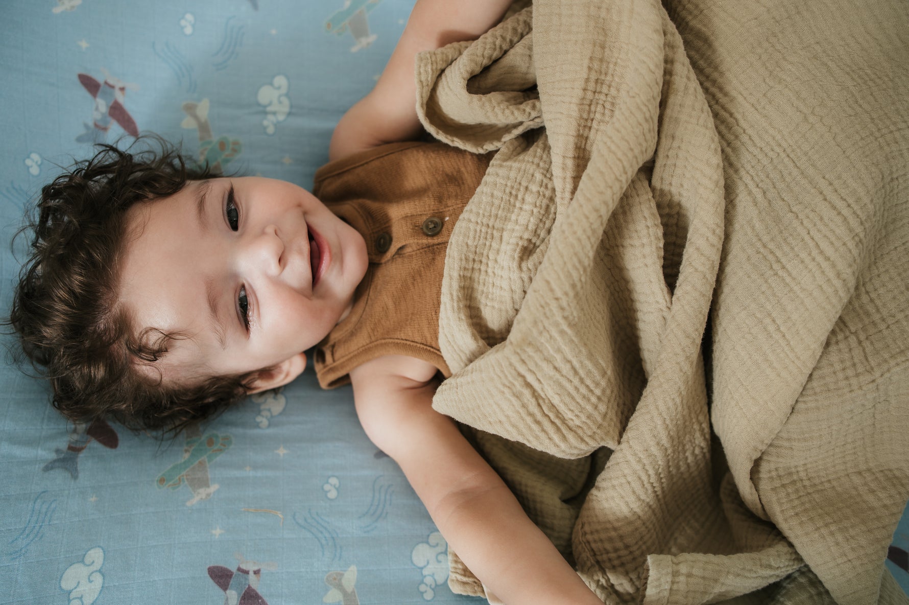 Child lying on a bed with a plane crib sheet with a blanket, looking up