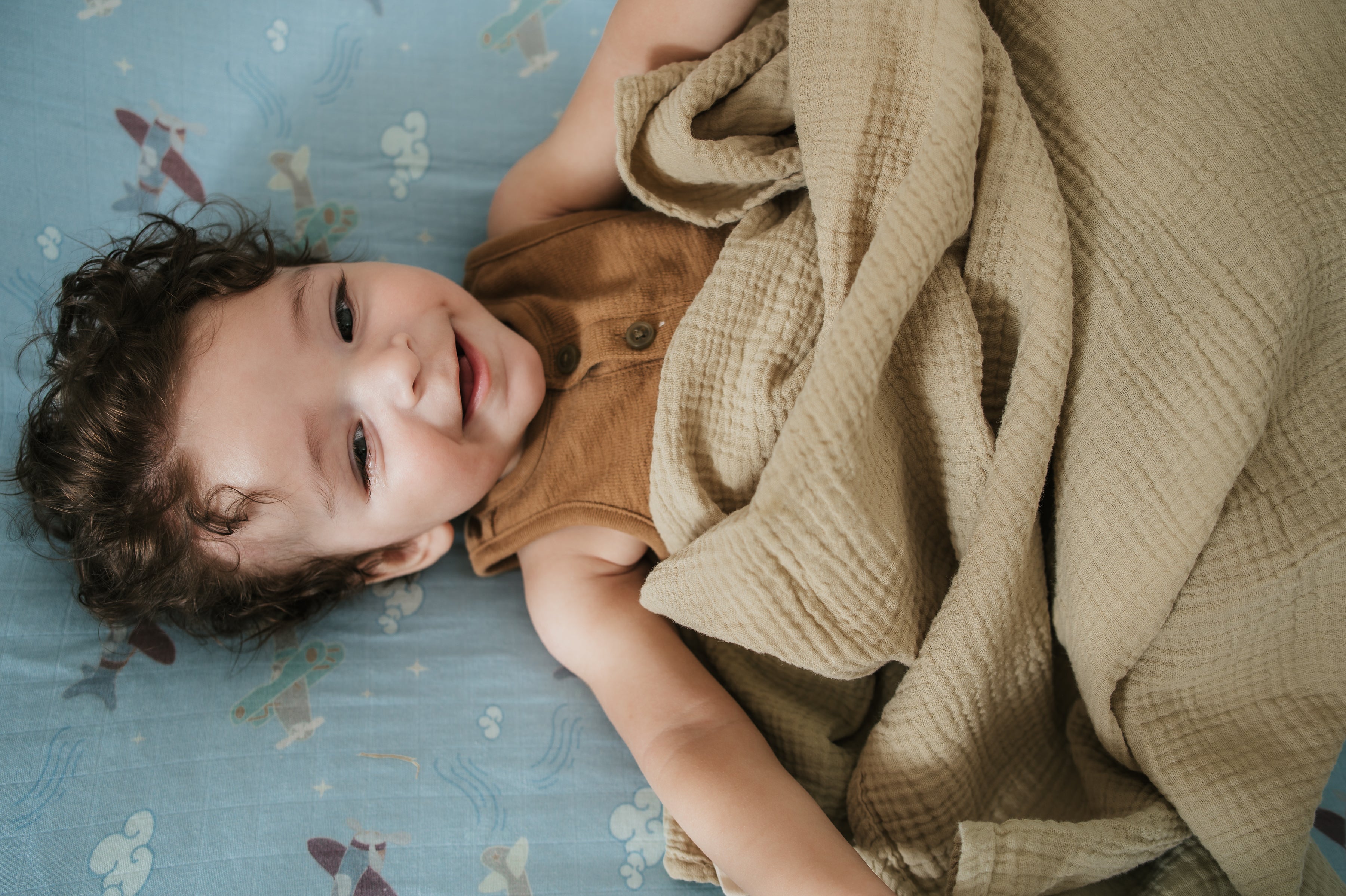 Child lying on a bed with a plane crib sheet with a blanket, looking up