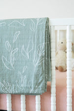 Green leaf-patterned blanket draped over a crib with a teddy bear inside.