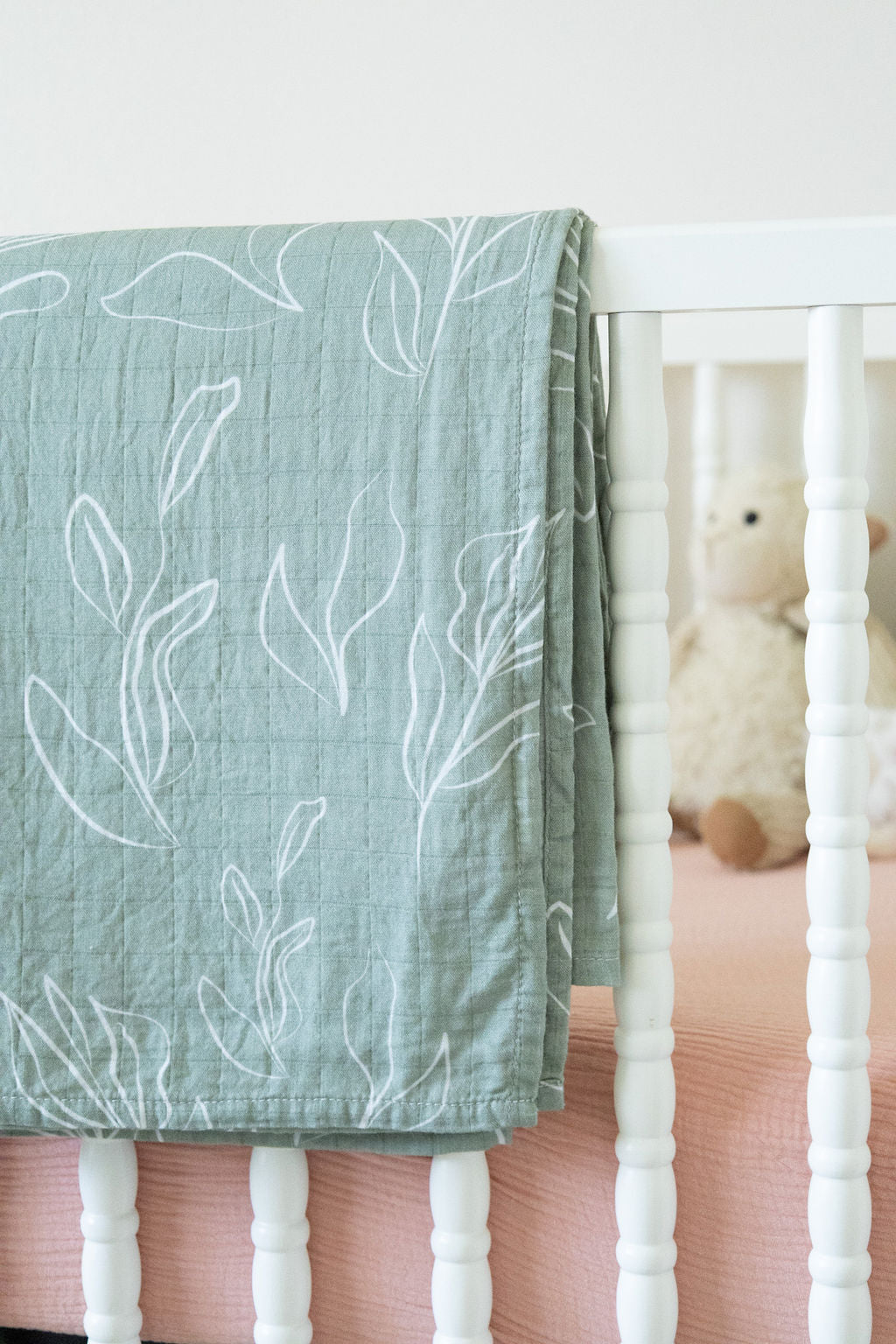 Green leaf-patterned blanket draped over a crib with a teddy bear inside.